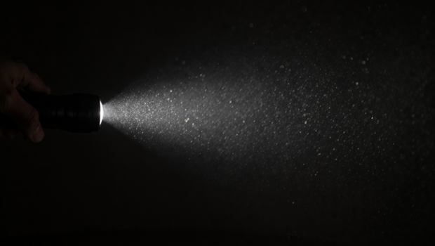 暗闇の中で懐中電灯を照射し、空中の微細な粒子が光を反射（散乱）して白く輝いている様子。散乱光方式において、粒子に当たって跳ね返る光（散乱光）を測定する仕組みを視覚化したイメージです。